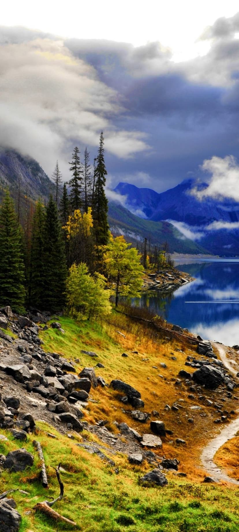 Misty mountain peaks loom over a reflective alpine lake with golden autumn foliage and evergreen trees on the shore.