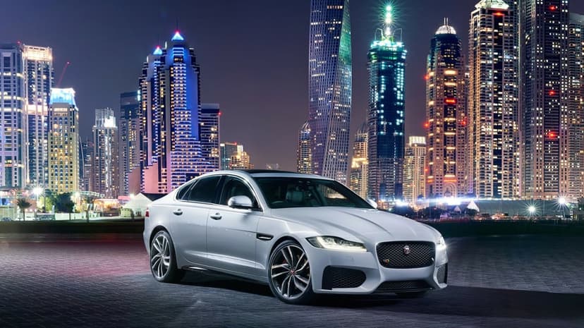 Silver luxury sedan parked on a dark plaza with illuminated skyscrapers and city lights in the night background.