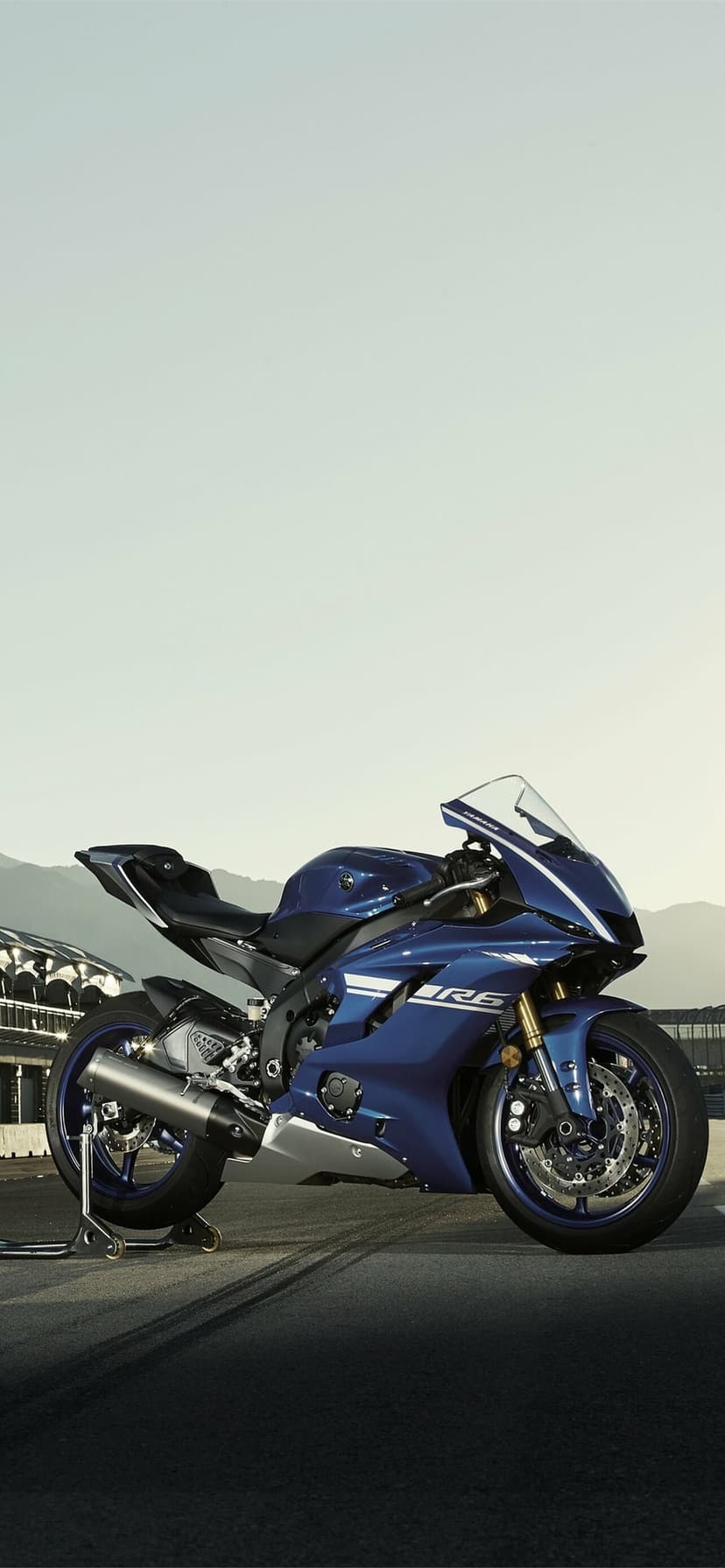 Blue Yamaha R6 sportbike parked on a track paddock stand against distant mountains and a soft sky.