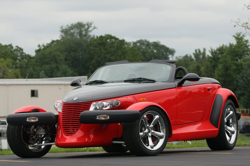 Red and black Plymouth Prowler with chrome wheels parked on asphalt under a cloudy sky with green trees.