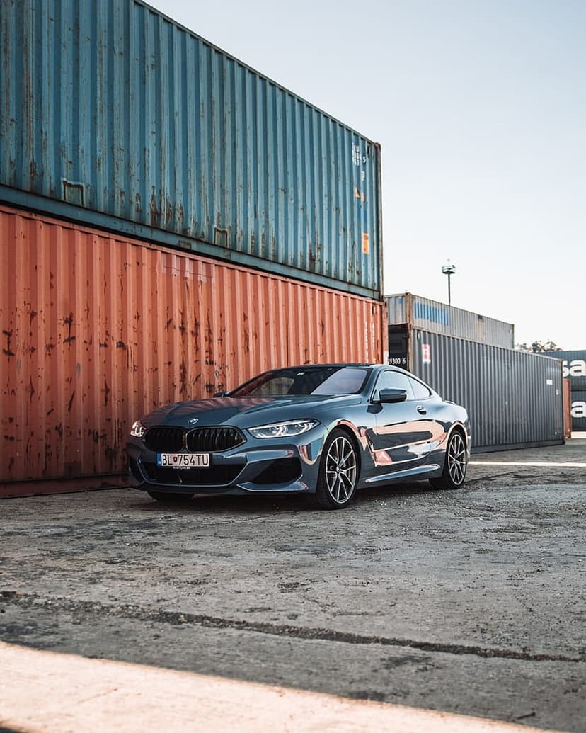 Dark blue BMW coupe parked next to orange and blue shipping containers on a sunlit concrete industrial lot.