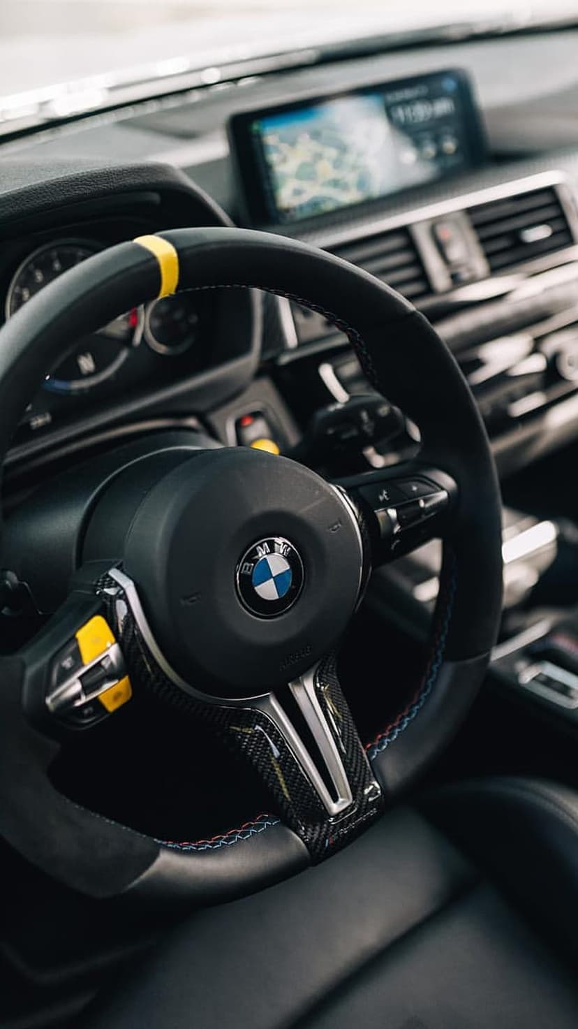 BMW M steering wheel with carbon fiber, yellow stripe, and tri-color stitching against a glowing dashboard.