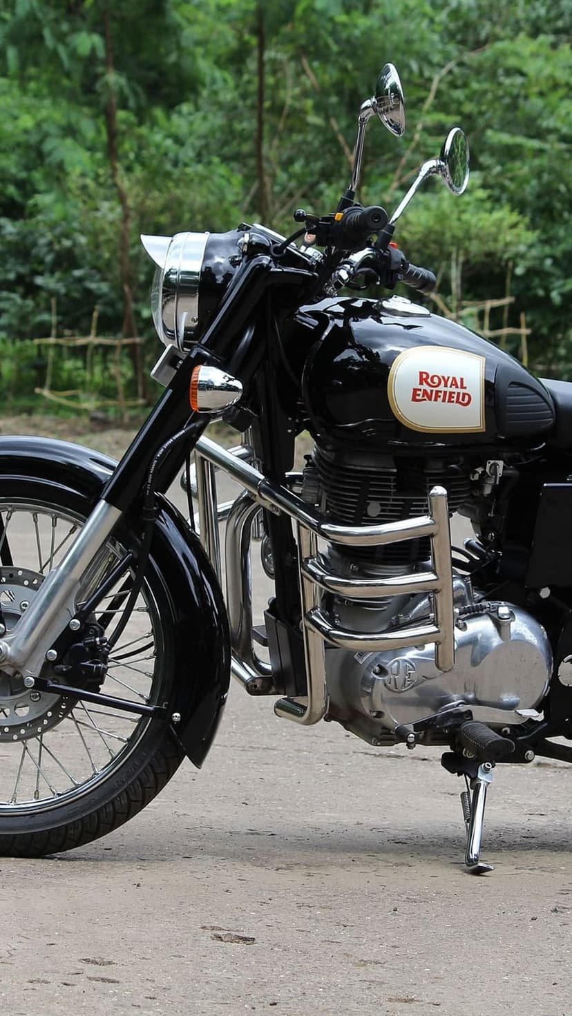 Black Royal Enfield motorcycle with chrome engine parts parked on a gravel road surrounded by green leaves.