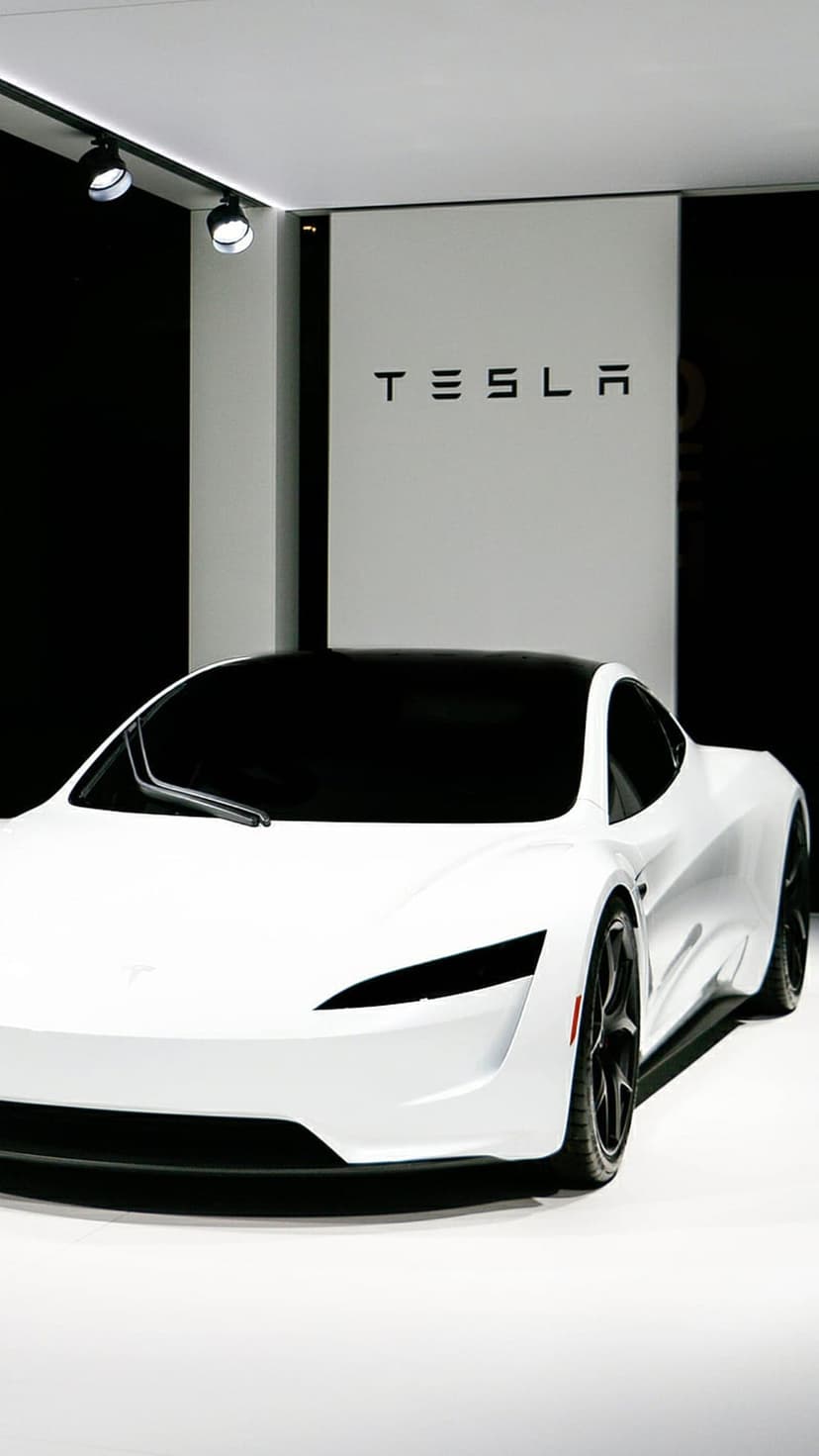 White Tesla sports car with a black roof parked in a showroom under spotlights in front of a branded backdrop.