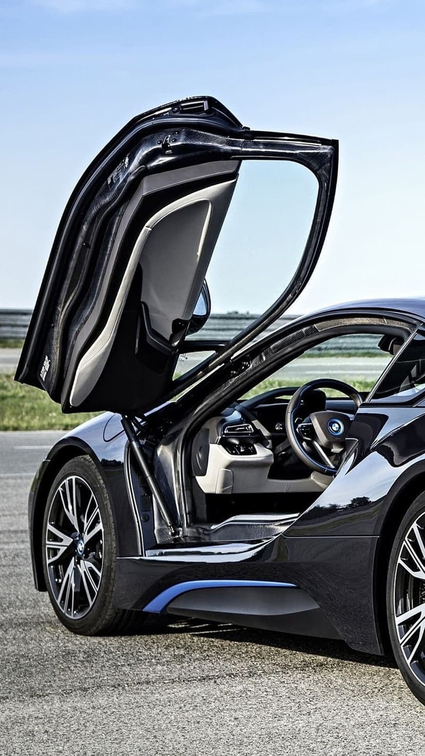 Black BMW i8 sports car with butterfly doors open on an asphalt road under a clear blue sky in bright daylight.