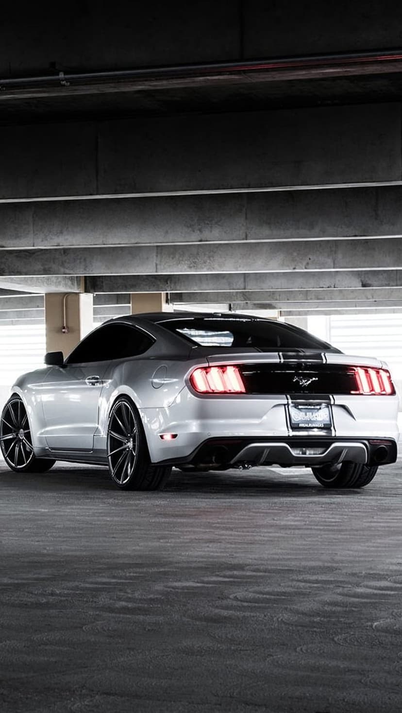 Silver Ford Mustang rear with black racing stripes and custom rims parked inside a dark concrete parking garage.