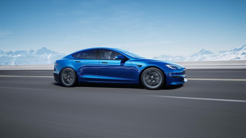 Blue Tesla electric car speeds down a wide mountain road with snow-covered peaks under a clear blue sky.