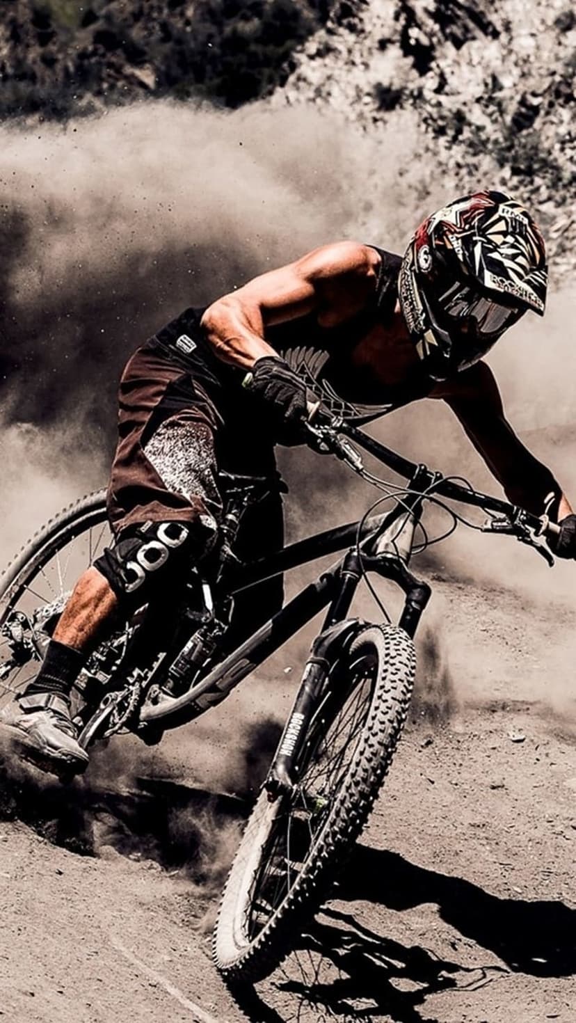 Mountain biker in full gear leans into a sharp dirt turn while kicking up a thick cloud of dust on a forest trail.