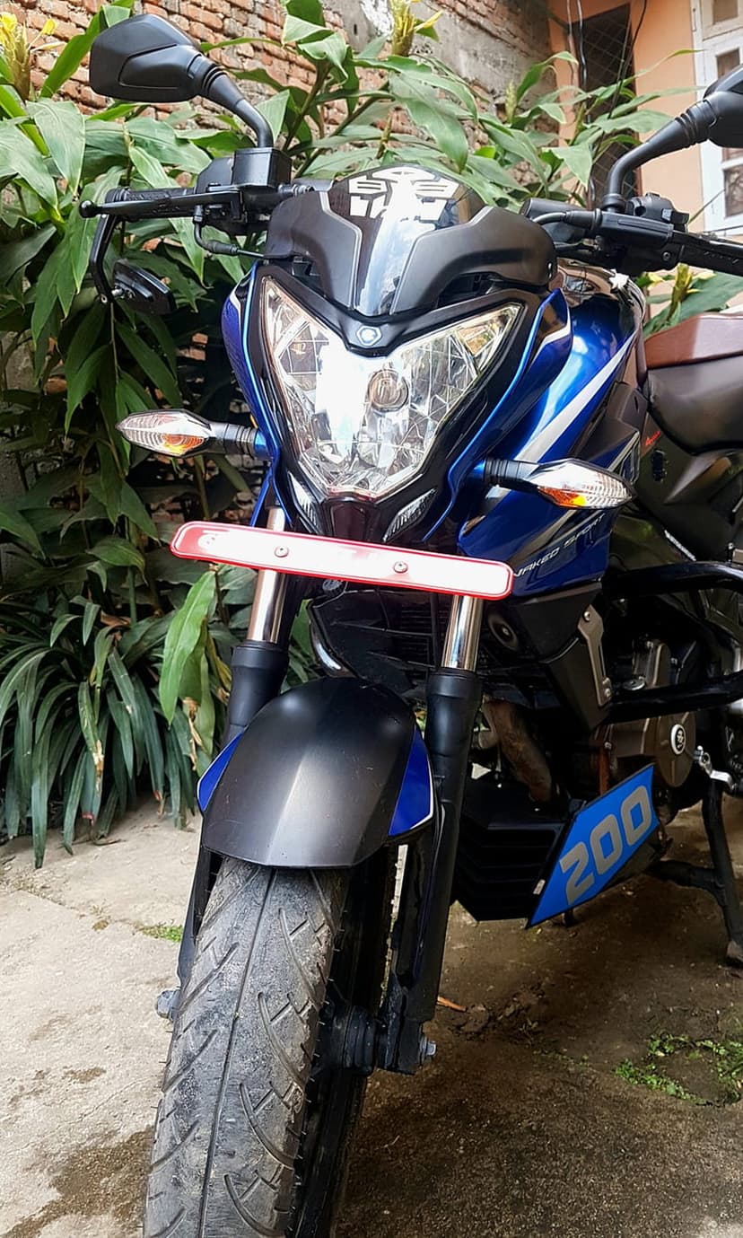 Blue Bajaj Pulsar 200 motorcycle showing dual headlights and handlebars against a green leafy background.