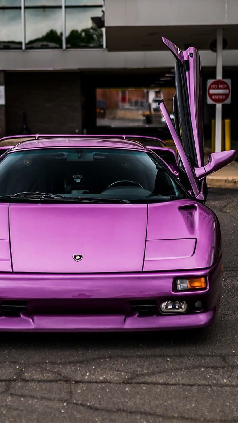Purple Lamborghini Diablo with open scissor doors parked on asphalt near a building with glass reflections.
