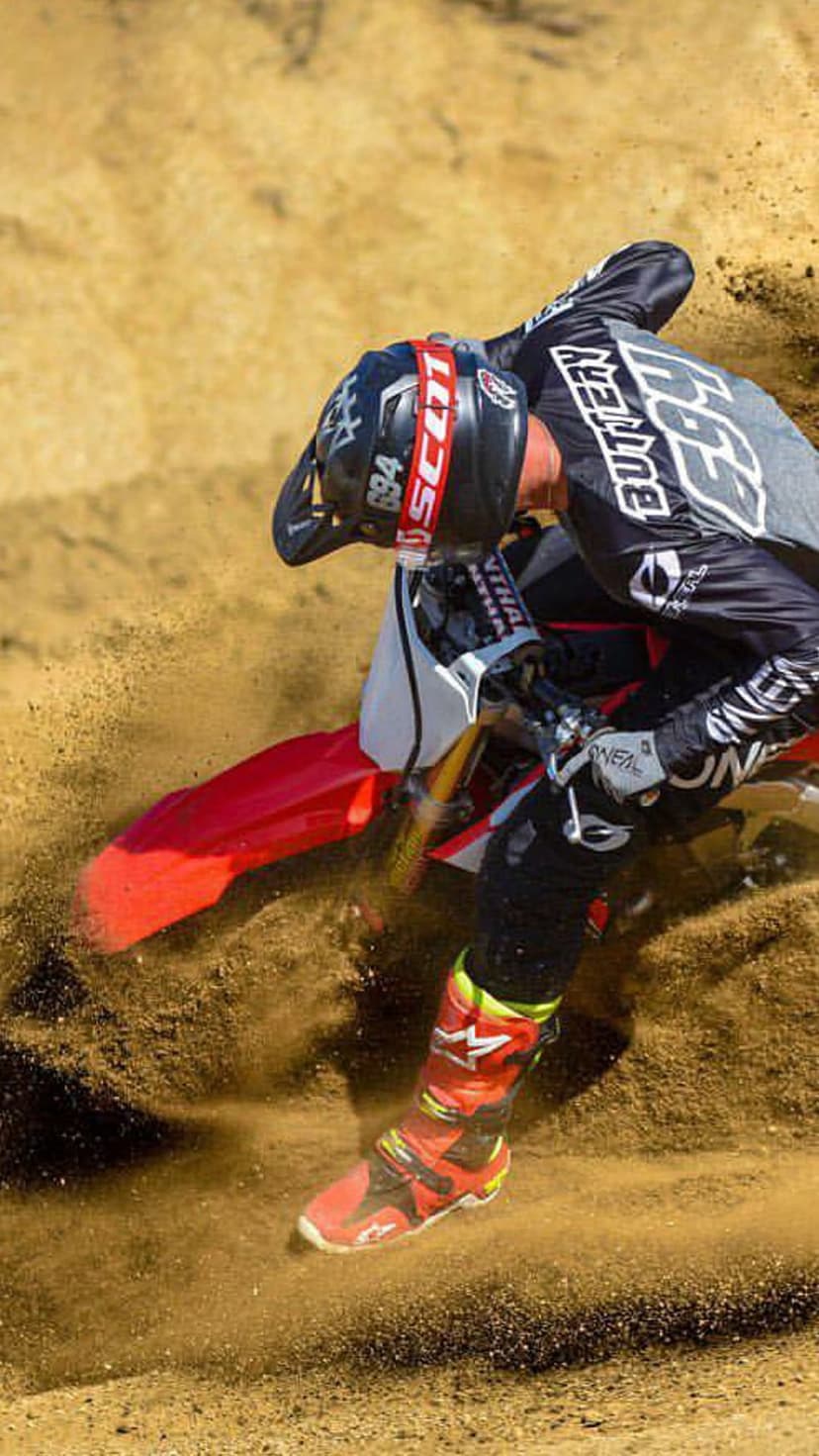 Motocross rider leaning into a sandy turn with a massive spray of dirt, wearing full racing gear and helmet.