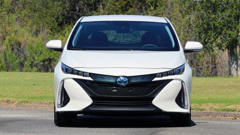 Front view of a white Toyota Prius Prime with LED headlights and blue emblem against a background of green trees.