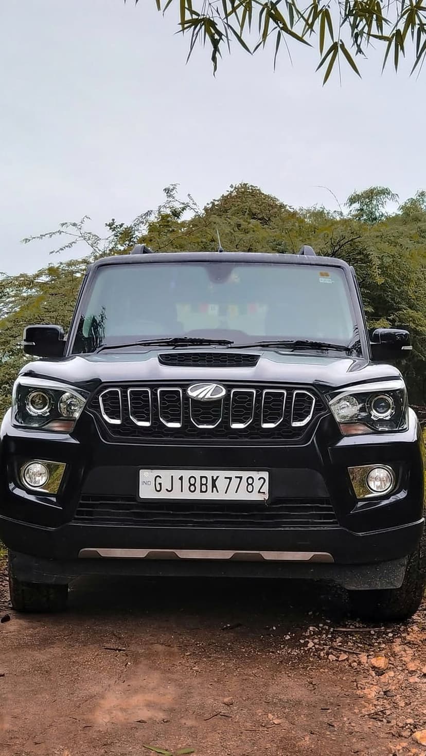 Black Mahindra Scorpio SUV with chrome grille on a dirt path surrounded by lush green trees and foliage.