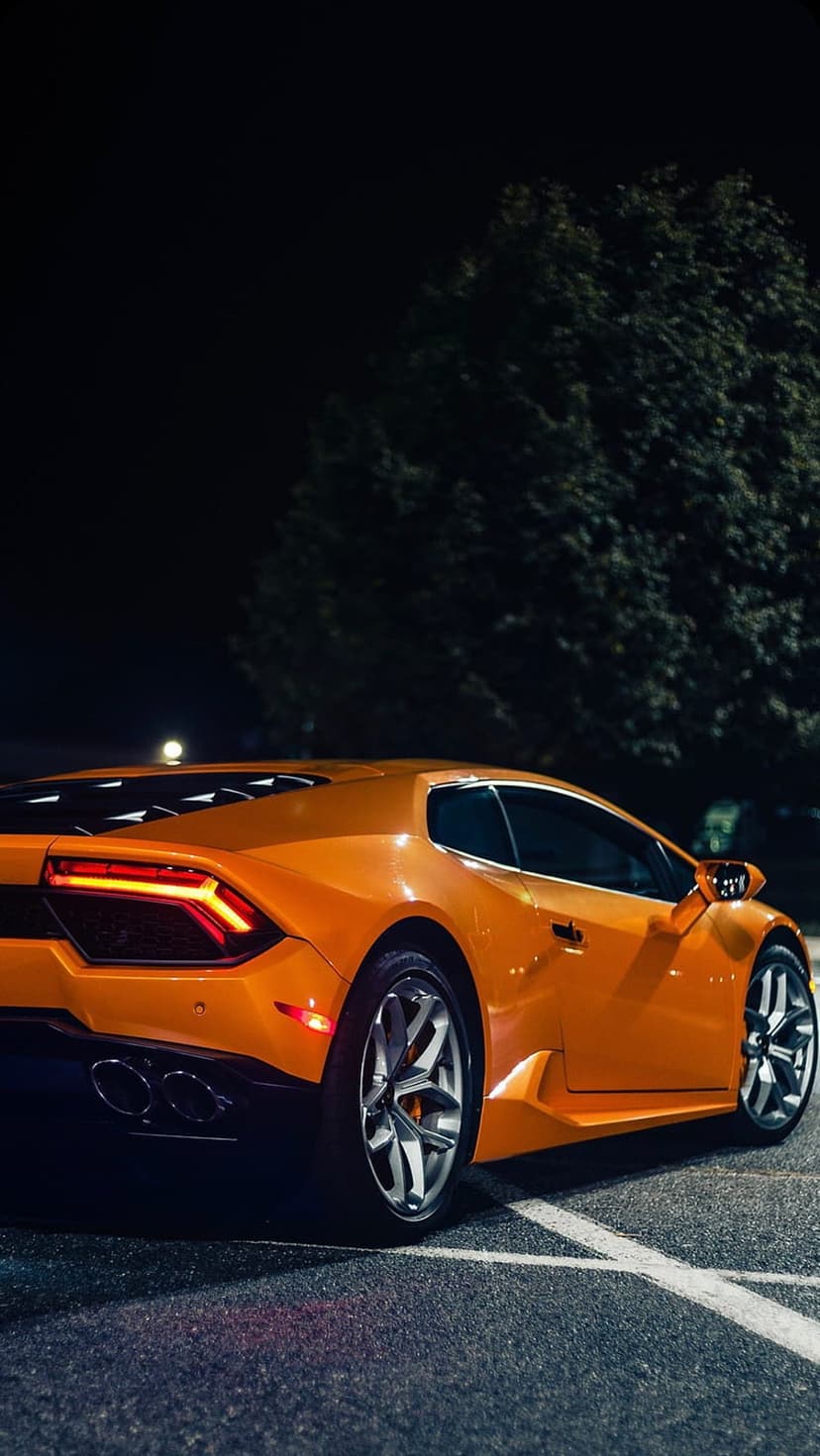 Orange Lamborghini Huracan with glowing taillights parked near a dark tree on asphalt under night lighting.