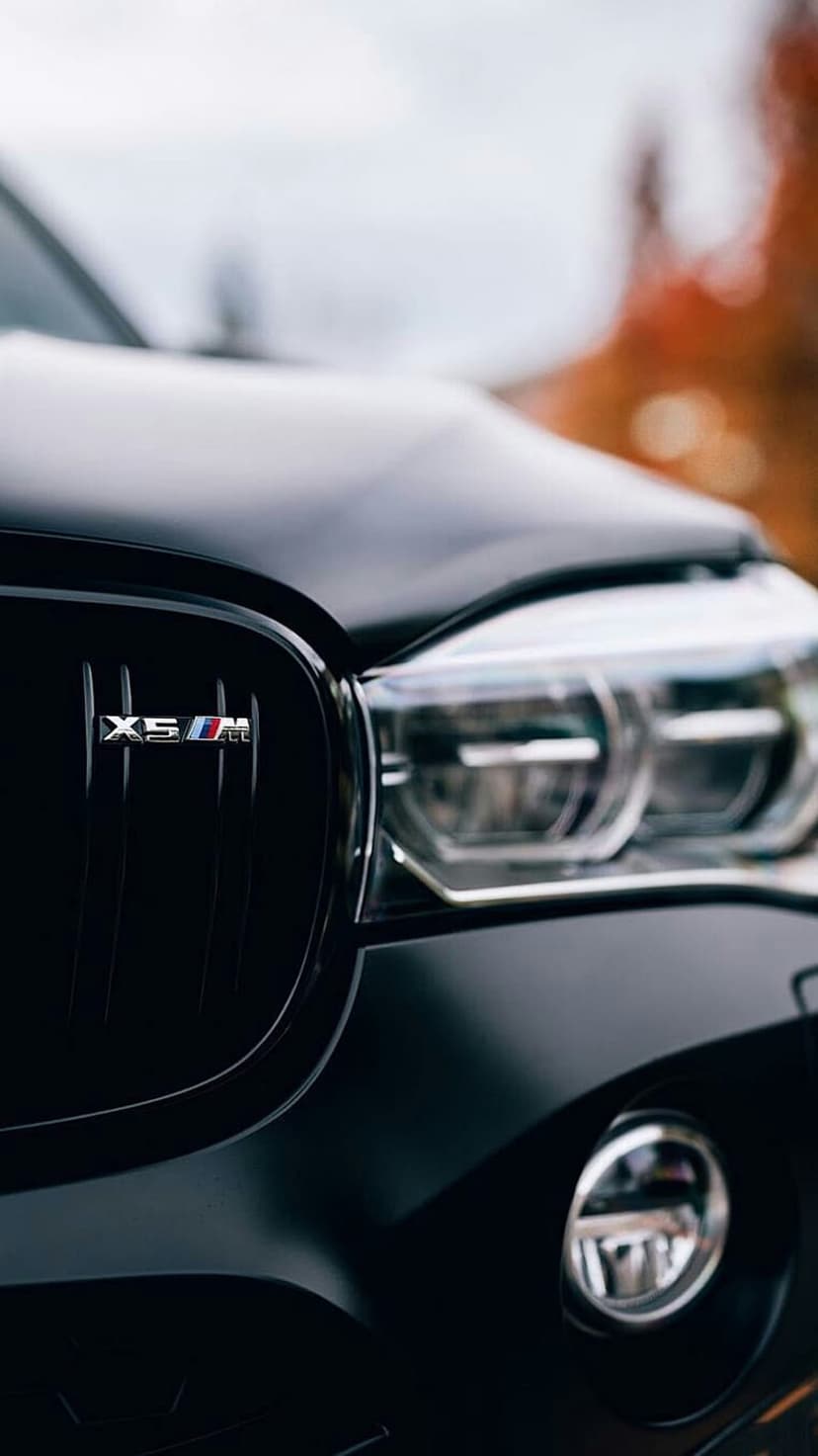 BMW X5M matte black front grille and LED headlights with an M-series badge against a blurred autumn background.