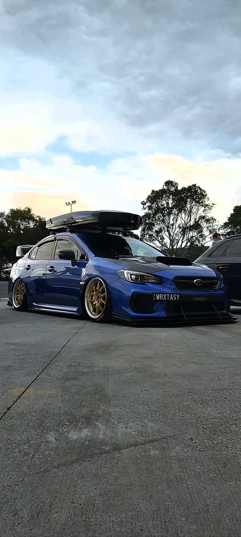 Blue Subaru WRX STI sedan featuring a black roof box and gold wheels parked on concrete under a cloudy sky.