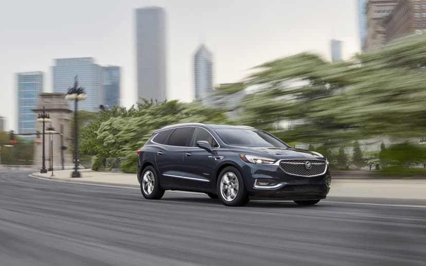 Dark grey Buick Enclave Avenir SUV driving on a city street with motion blurred trees and skyscrapers.