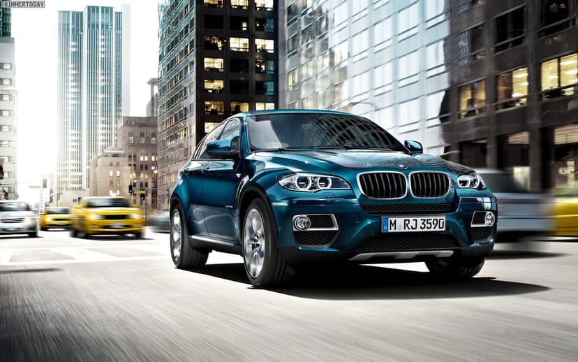 Teal BMW X6 luxury SUV driving on a city street with motion blur and tall glass skyscrapers in the background.
