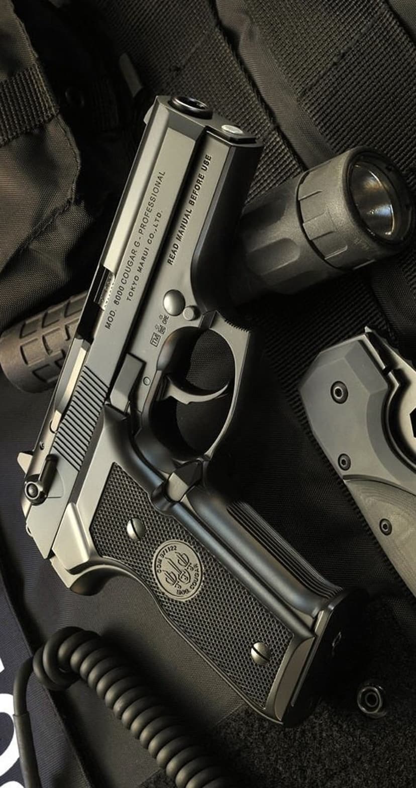 Beretta 8000 Cougar F pistol beside a folding knife and flashlight on textured dark fabric in a close-up shot.