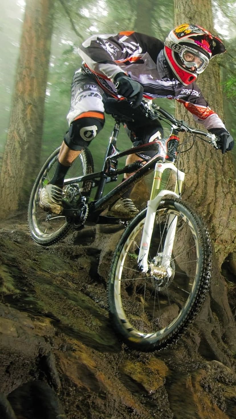 Mountain biker wearing a full-face helmet descends a steep, rocky forest trail on a full-suspension bicycle.