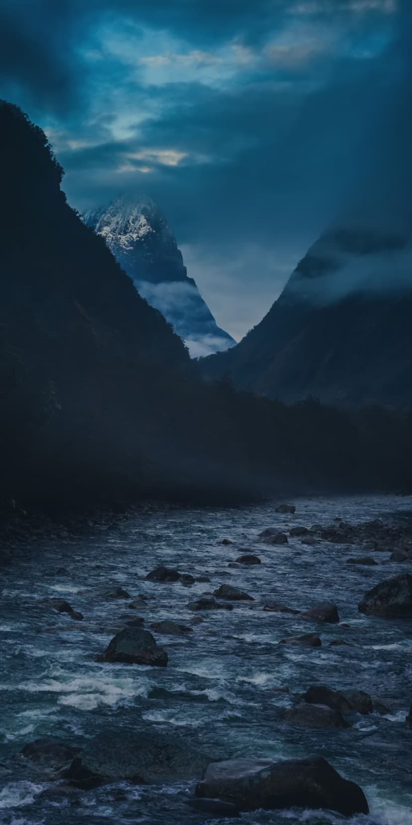 Rugged mountains frame a turbulent river under misty skies with a distant snow-capped peak in New Zealand.
