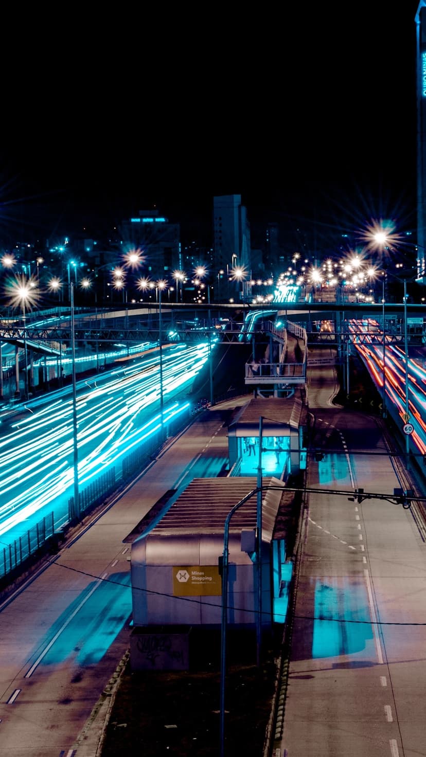 Luminous blue and red car light trails streak down a dark urban street lined with glowing modern skyscrapers.