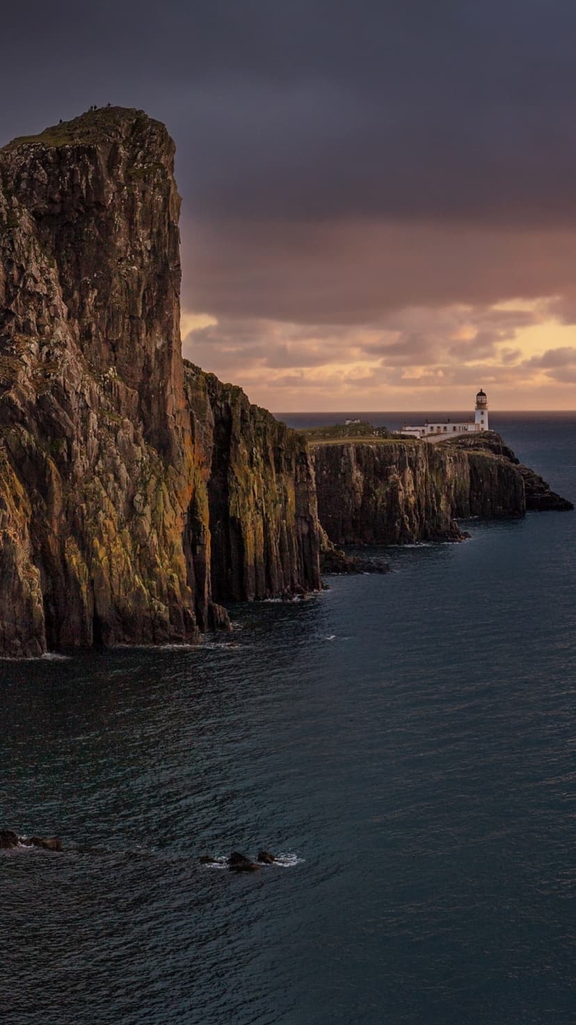 Neist Point Lighthouse perched on steep cliffs overlooking the dark Atlantic Ocean under a stormy twilight sky.