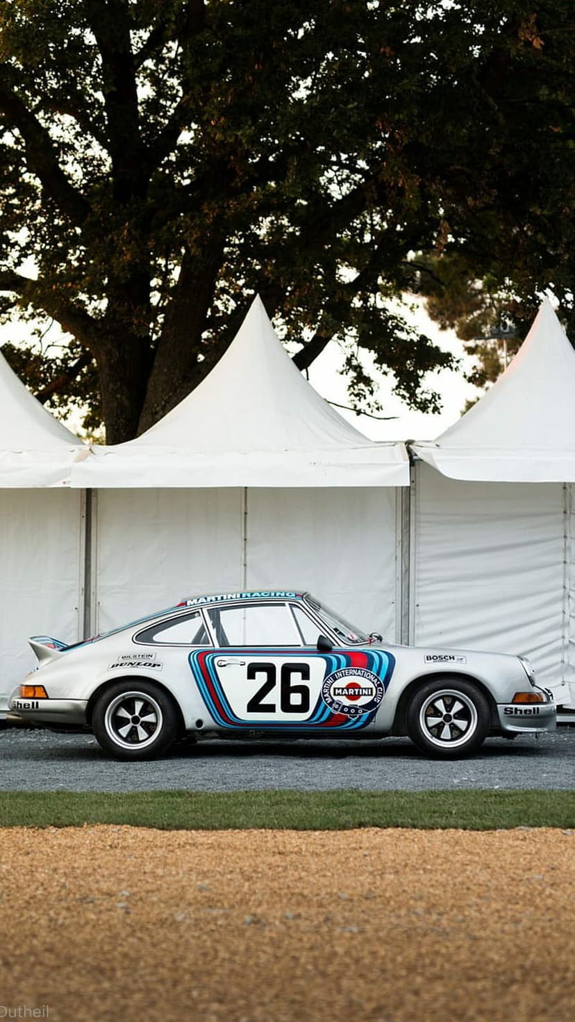 Silver Porsche 911 race car with Martini livery and number 26 parked outdoors under a tree at a car show.