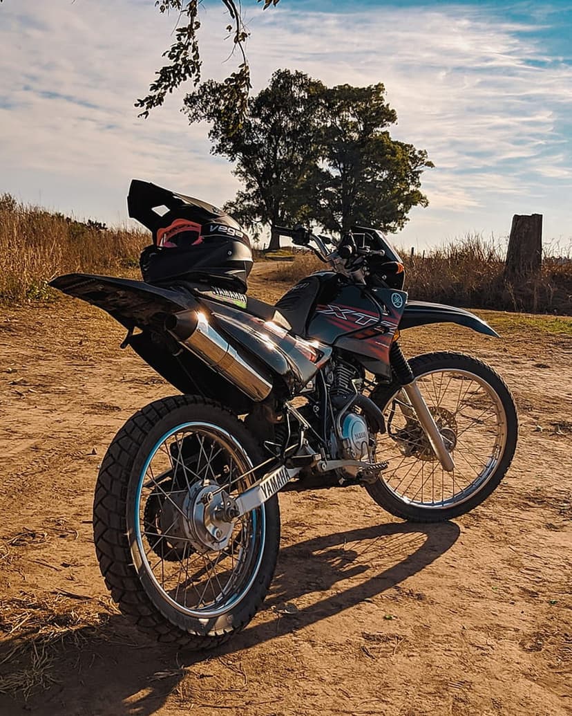Yamaha dirt bike with a helmet on the seat parked on a sandy trail under a cloudy sky and green tree branches.