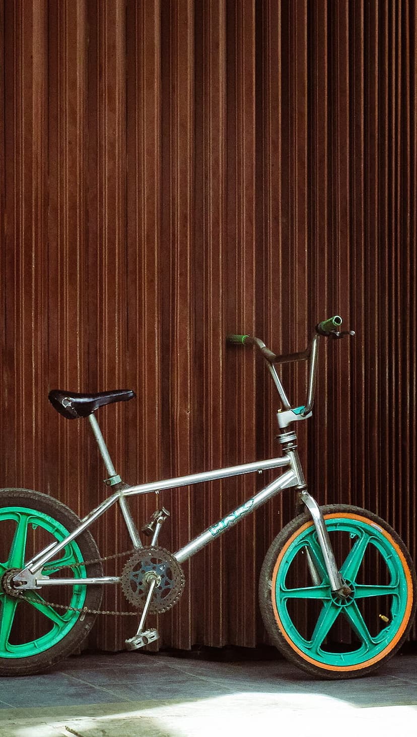 Silver BMX bicycle with teal five-spoke wheels and orange rims parked against a brown corrugated metal rollup door.