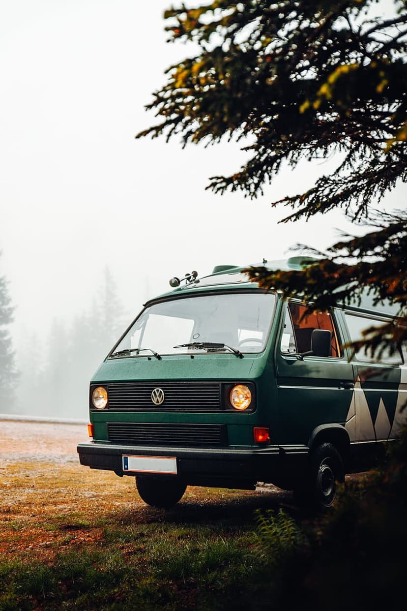Green vintage Volkswagen van parked on grass surrounded by tall trees and thick white fog in a secluded forest.
