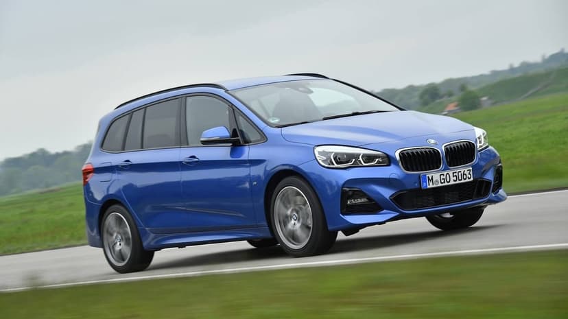 Blue BMW Gran Tourer driving on a track with motion-blurred wheels against a green hillside background.