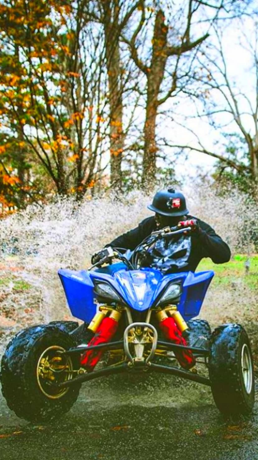 Person in a black helmet rides a blue ATV with gold rims through splashing mud and water in an autumn forest.