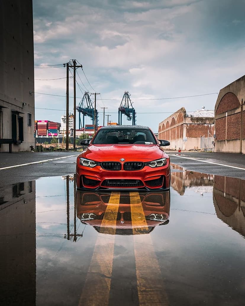 Red BMW M4 sports car reflected in a puddle on wet asphalt with industrial cranes under a cloudy grey sky.