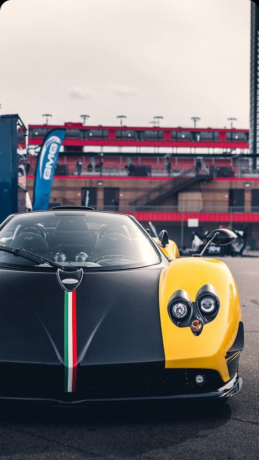 Yellow Pagani supercar with carbon fiber bodywork and Italian flag stripe at a race track under daylight.