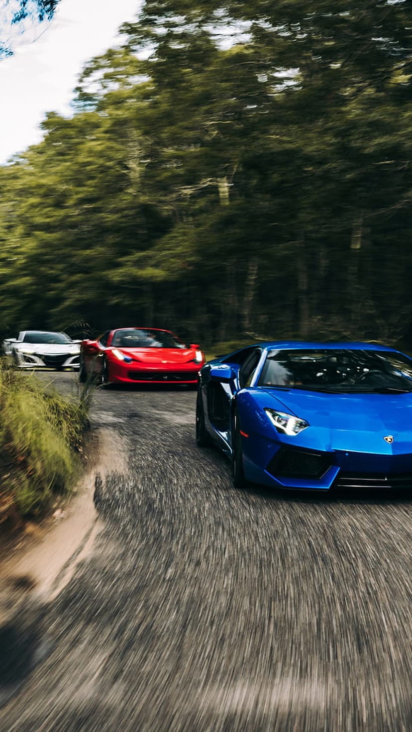 Blue Lamborghini, red Ferrari, and white Acura NSX speed along a winding forest road with motion blur.