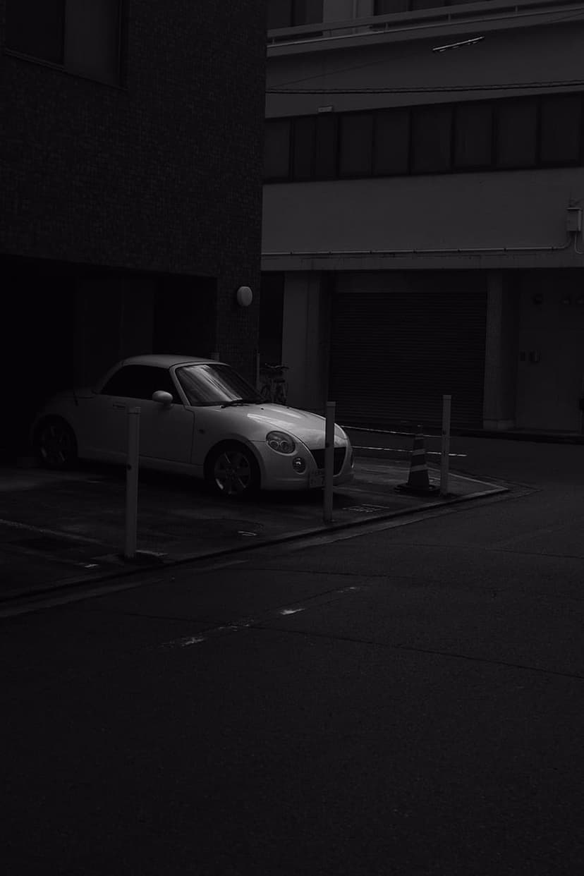 White convertible car parked between dark brick walls in a monochrome urban alleyway with high-contrast lighting.