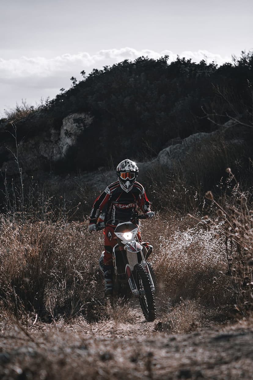 Motocross rider in full gear navigates a dirt bike through tall dry grass and rocky hills under a cloudy sky.