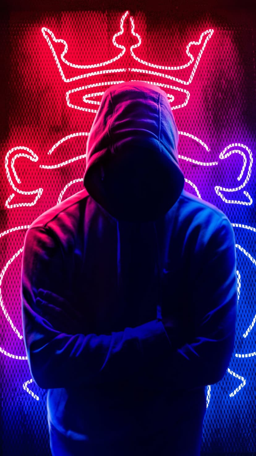 Hooded person with crossed arms standing before a glowing red and blue neon crown within a dark urban setting.