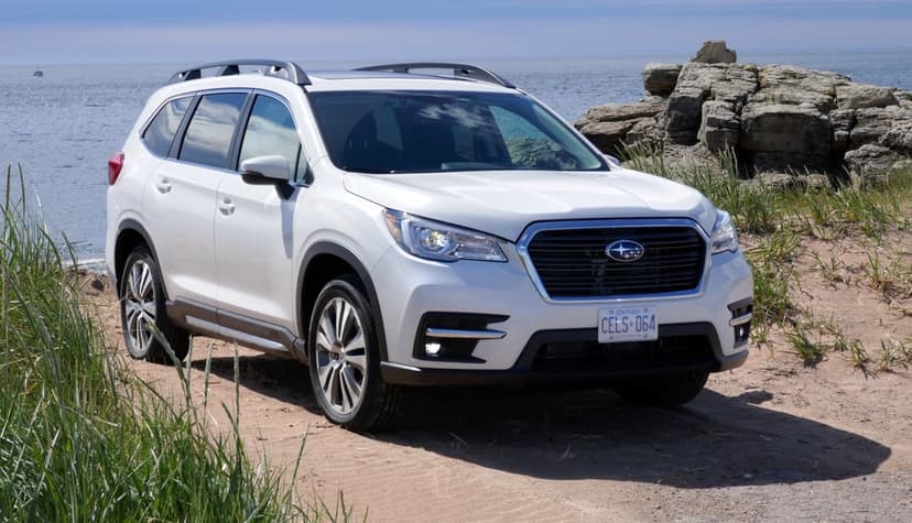 White Subaru Ascent SUV parked on a sandy coastal path with ocean waves and rocky cliffs in the background.