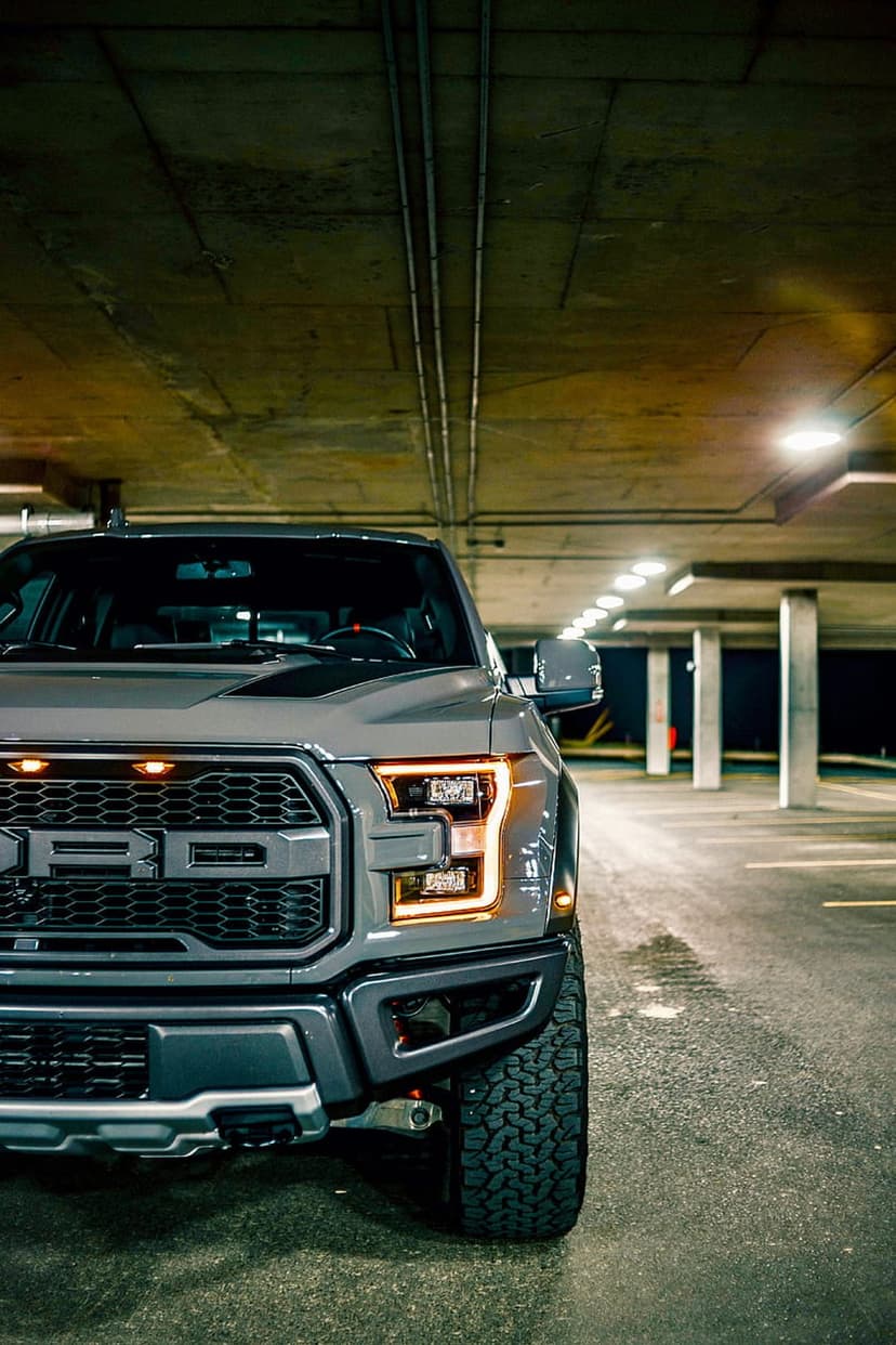 Grey Ford Raptor truck with glowing amber lights parked in a dark industrial garage with concrete ceilings.