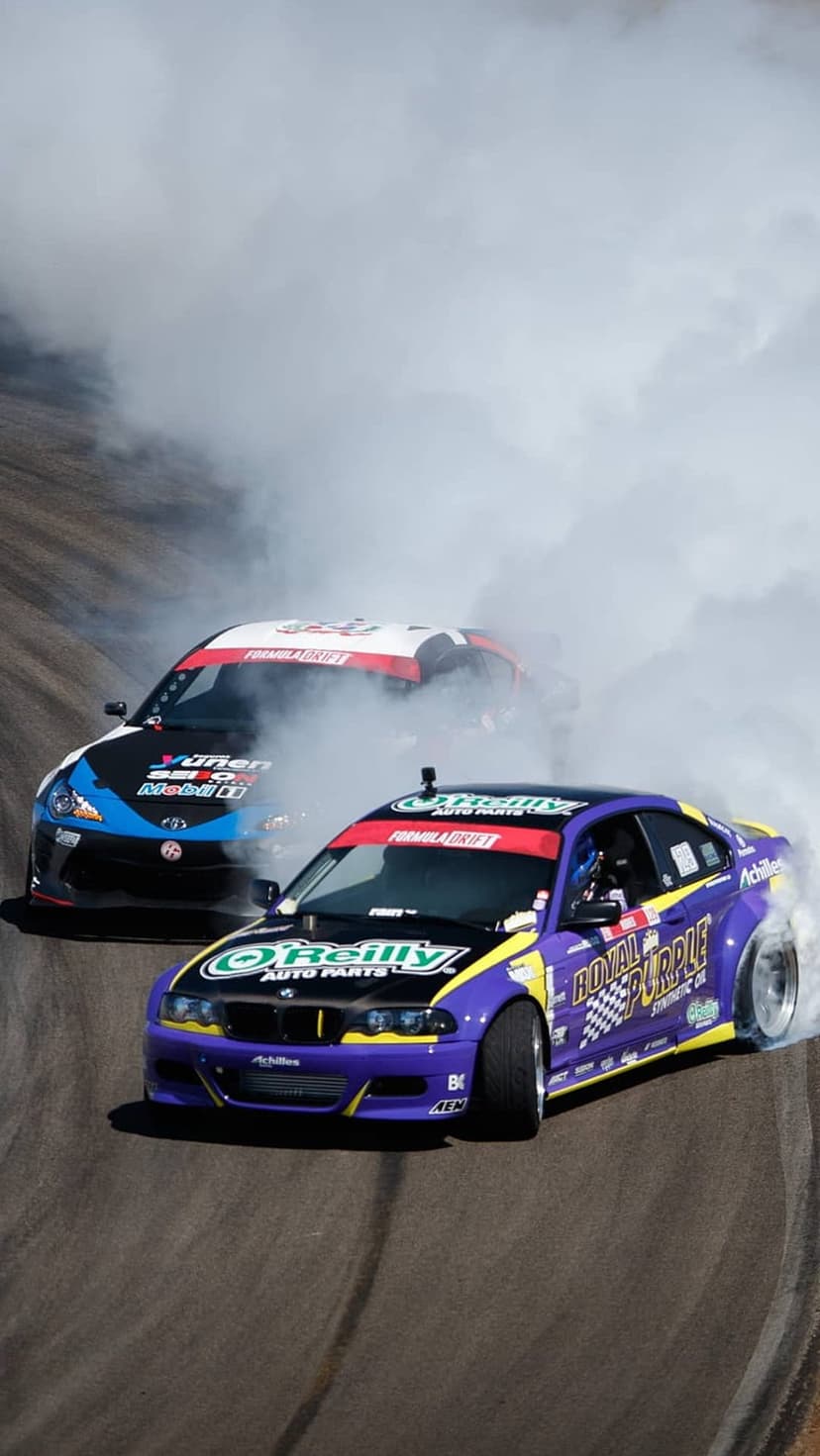Purple and blue drift cars slide sideways on a racing circuit amidst clouds of white smoke for a phone screen.