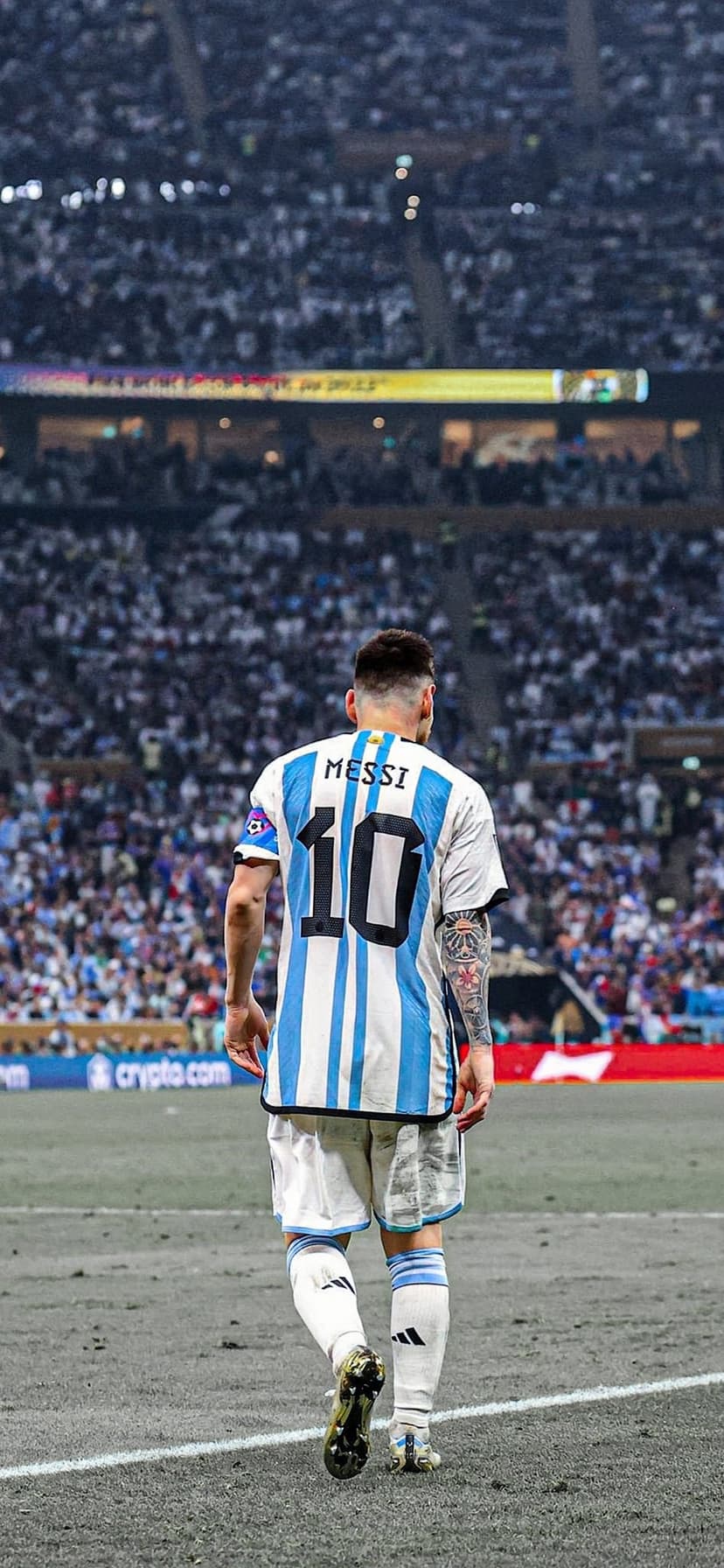 Lionel Messi walks away on a grass pitch wearing a striped Argentina jersey with number 10 in a packed stadium.