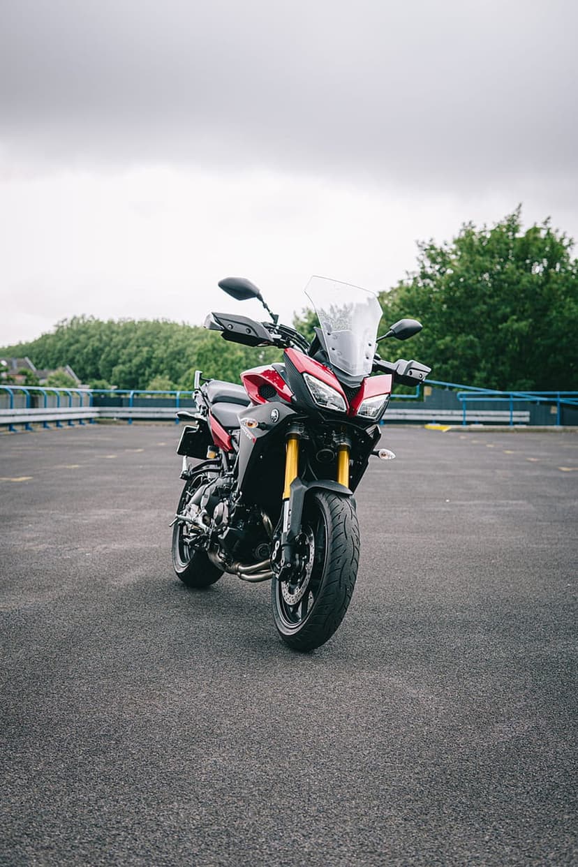 Red Yamaha motorcycle with yellow forks parked on asphalt against lush green trees and a cloudy gray sky.