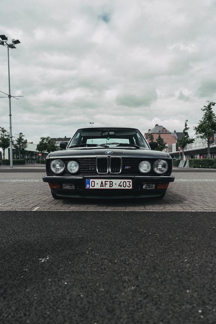 BMW E28 M535i sedan with a black finish and classic round headlights parked on a gray cobblestone street.