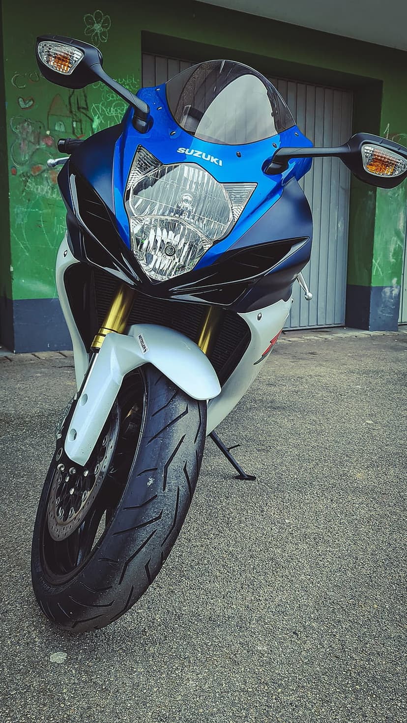 Suzuki GSX-R sportbike front with blue fairings, golden suspension, and textured tires against a graffiti wall.