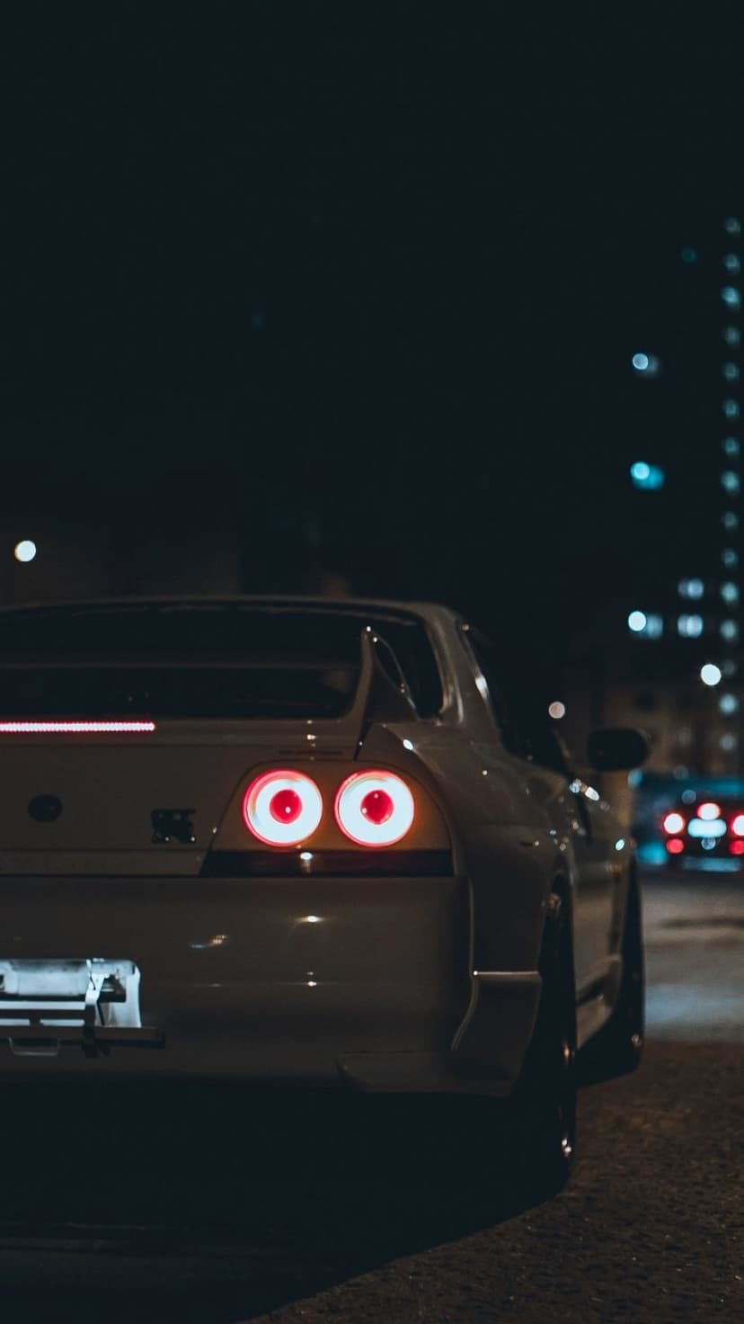 Rear view of a white Nissan Skyline R33 GT-R at night with glowing red round taillights and city bokeh.