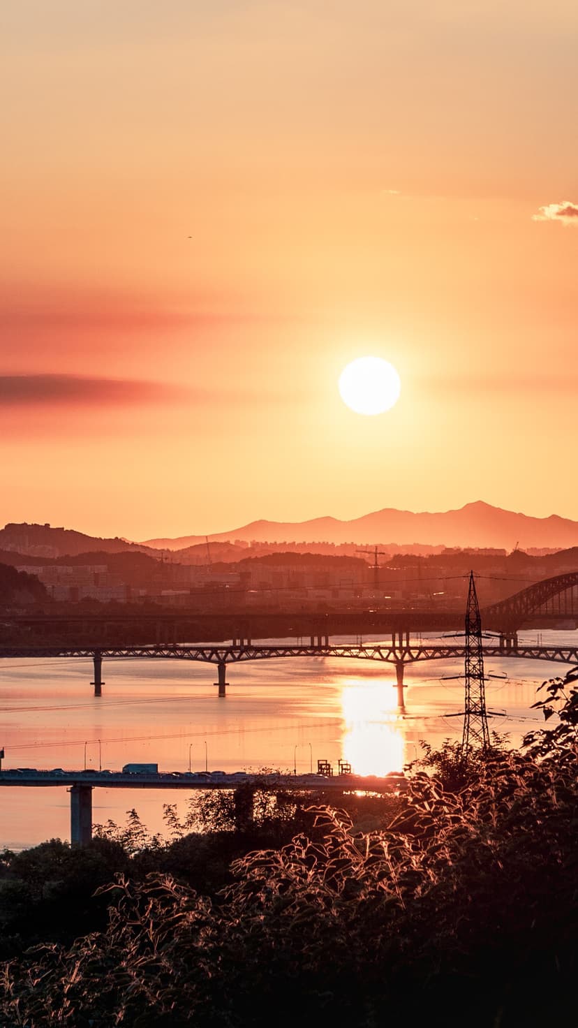 Orange sun reflects on the Han River with city bridges and hills, captured as a vertical phone background.