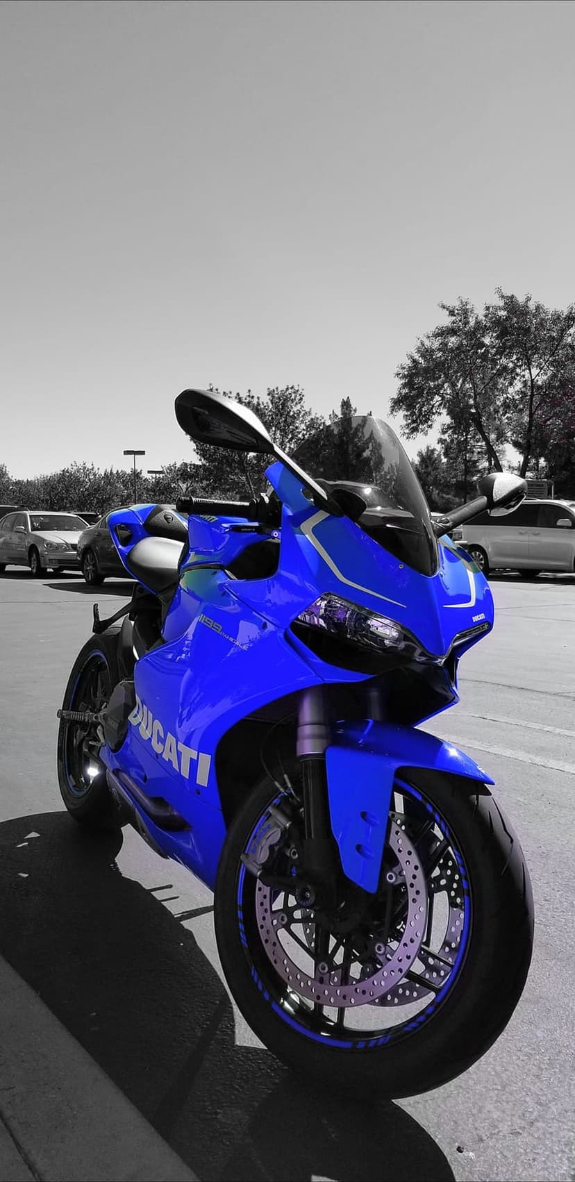 Blue Ducati motorcycle parked in a grayscale parking lot with trees and cars in the desaturated background.