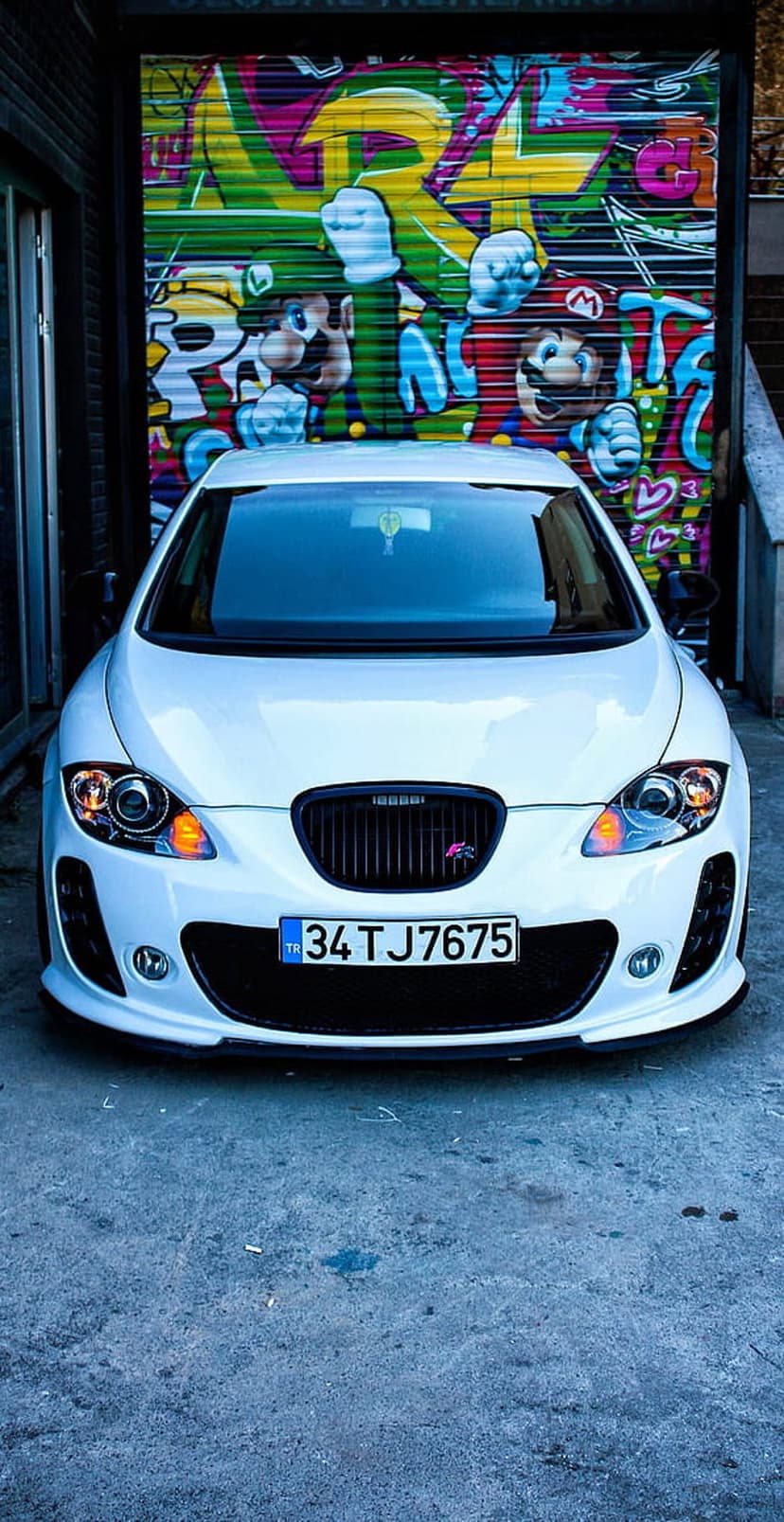 White car front view parked before a colorful garage door with painted Mario characters and vibrant urban art.